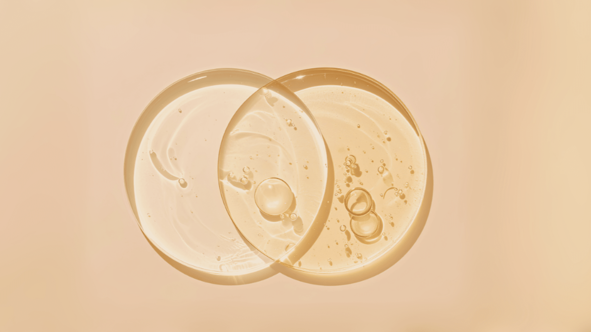 Two overlapping transparent discs filled with clear and golden liquid, showing small air bubbles and smooth textures, set against a soft pastel background with gentle light reflections
