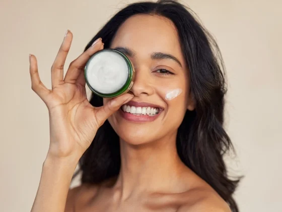 Smiling woman with dark hair holding an open jar of face cream over one eye, with a small amount of cream applied to her cheek, against a neutral background.