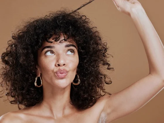 Woman with curly hair playfully holding up a single curl, looking upward with pursed lips, against a warm neutral background.
